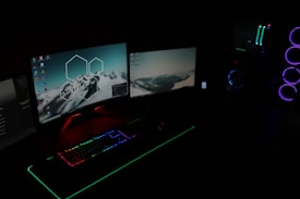 A dark room illuminated by two computer monitors displaying desktop backgrounds with mountain images. A gaming setup features a keyboard with RGB lighting and a tower with LED fans glowing in blue and purple.