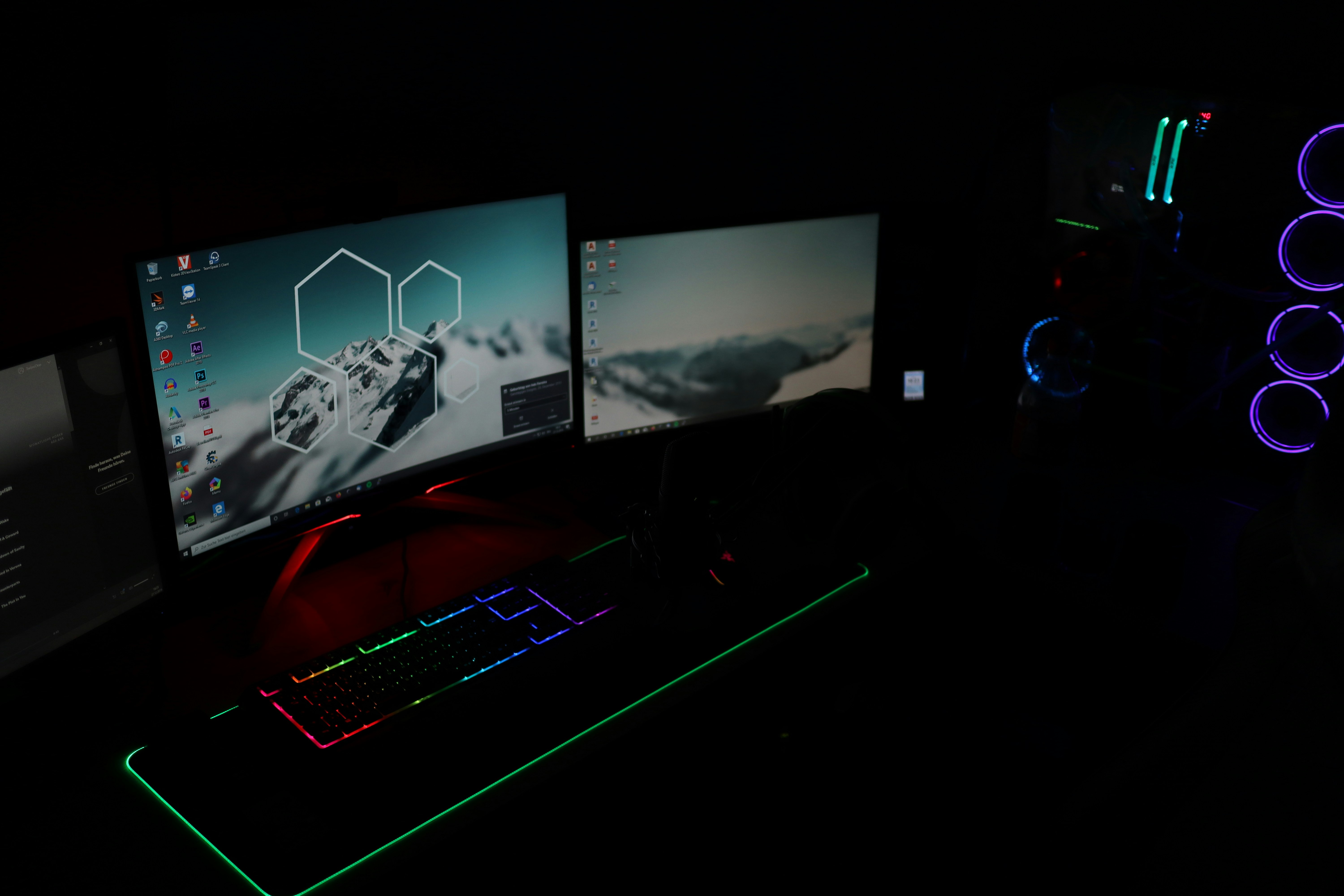 A dark room illuminated by two computer monitors displaying desktop backgrounds with mountain images. A gaming setup features a keyboard with RGB lighting and a tower with LED fans glowing in blue and purple.