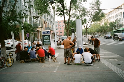 A group of people gather around on the sidewalk, with some sitting on small stools while others stand. They're concentrated on a central activity, possibly a game or discussion. The setting is urban, with tall buildings, parked cars, bicycles, and trees lining the street.
