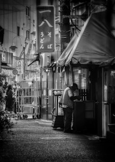 A narrow urban alleyway features a person standing near the entrance of a small shop with Japanese signage. The scene includes hanging signs and visible wires overhead, and the overall environment suggests a bustling street. A few people can be seen in the background near a stack of metal carts.