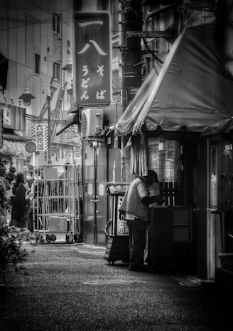 A narrow urban alleyway features a person standing near the entrance of a small shop with Japanese signage. The scene includes hanging signs and visible wires overhead, and the overall environment suggests a bustling street. A few people can be seen in the background near a stack of metal carts.