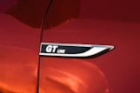 A close-up of a shiny, metallic badge with the text 'GT Line' on a surface with a glossy red paint finish. The badge is bordered by a sleek silver trim.