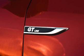 A close-up of a shiny, metallic badge with the text 'GT Line' on a surface with a glossy red paint finish. The badge is bordered by a sleek silver trim.
