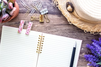 A rustic wooden desk with a notebook, pen, and a signature handcrafted hat resting beside fresh wildflowers.