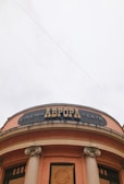 The image depicts the facade of a building with a round structure and two large columns. The building has an orange hue, and a black sign with the word 'CINEMA' in large letters is visible. Above the entrance, there are decorative elements, including a marquee with an emblem in the center. The sky is overcast, giving a neutral backdrop.