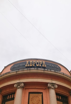 The image depicts the facade of a building with a round structure and two large columns. The building has an orange hue, and a black sign with the word 'CINEMA' in large letters is visible. Above the entrance, there are decorative elements, including a marquee with an emblem in the center. The sky is overcast, giving a neutral backdrop.