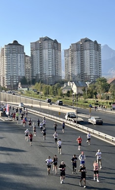A group of people is running on a closed-off road, possibly participating in a marathon or race. The background features several tall residential buildings and a clear sky, with a mountain visible in the distance. Some spectators and cars are seen on the roadside.