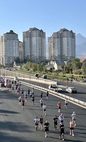A group of people is running on a closed-off road, possibly participating in a marathon or race. The background features several tall residential buildings and a clear sky, with a mountain visible in the distance. Some spectators and cars are seen on the roadside.