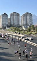 A group of people is running on a closed-off road, possibly participating in a marathon or race. The background features several tall residential buildings and a clear sky, with a mountain visible in the distance. Some spectators and cars are seen on the roadside.