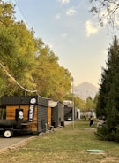 A row of food stalls, possibly made from shipping containers, are set in a park-like area. Trees line the path, and a mountain is visible in the distance. People are sitting and walking, suggesting a casual and relaxed environment.