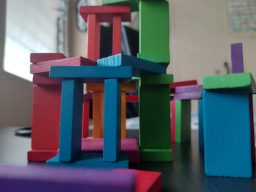 A collection of colorful wooden blocks is stacked in a playful and abstract manner, forming a miniature structure. The blocks vary in shape and color, creating a dynamic and vibrant arrangement.