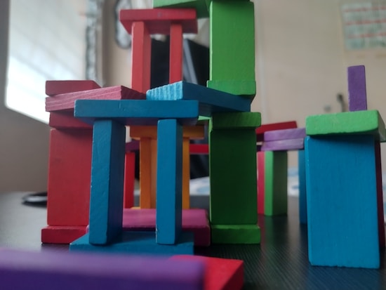 A collection of colorful wooden blocks is stacked in a playful and abstract manner, forming a miniature structure. The blocks vary in shape and color, creating a dynamic and vibrant arrangement.