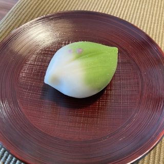 A beautifully crafted wagashi, traditional Japanese confectionery, shaped like a leaf. The sweet features a soft gradient of green to white with two delicate pink dots near the center, suggesting the presence of cherry blossoms or another floral detail. It is placed on a textured, dark red wooden plate with visible grain lines, against a woven beige placemat.