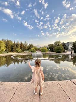 A young child wearing a light-colored dress stands on a stone platform facing a large circular fountain. The fountain is surrounded by water, reflecting the sky and trees. The sky is partly cloudy with a blue hue, and trees fill the background.
