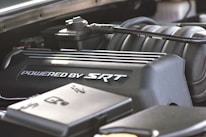 A close-up view of a car engine component, prominently featuring a black engine cover with the text 'Powered by SRT.' Various other engine parts are visible surrounding the cover, including tubes and metal components.