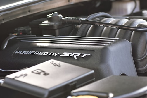 A close-up view of a car engine component, prominently featuring a black engine cover with the text 'Powered by SRT.' Various other engine parts are visible surrounding the cover, including tubes and metal components.