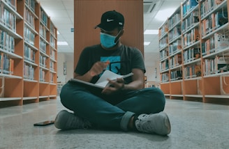 A person wearing a face mask and a cap is sitting cross-legged on the floor of a library. They are holding a notebook, seemingly engaged in writing or sketching, surrounded by bookshelves filled with colorful books.