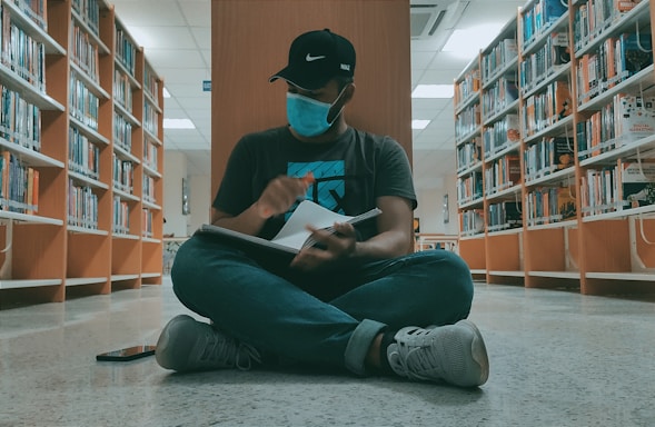 A person wearing a face mask and a cap is sitting cross-legged on the floor of a library. They are holding a notebook, seemingly engaged in writing or sketching, surrounded by bookshelves filled with colorful books.