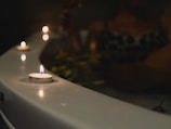 Several small candles are lit and placed along the edge of a bathtub in a dimly lit setting. A person appears to be relaxing in the tub, surrounded by floating flower petals.