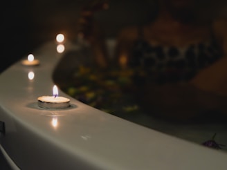 Several small candles are lit and placed along the edge of a bathtub in a dimly lit setting. A person appears to be relaxing in the tub, surrounded by floating flower petals.