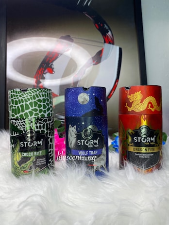 Three distinctively designed deodorant cans are placed on a fluffy white surface. The can on the left has a green and white crocodile skin pattern with the label 'CROCO BITE'. The middle can features a starry night sky with a wolf howling at the moon, labeled 'WOLF TRAP'. The can on the right showcases a red and gold dragon motif, labeled 'DRAGON FIRE'. In the background, there appears to be abstract art with swirling dark and red patterns.