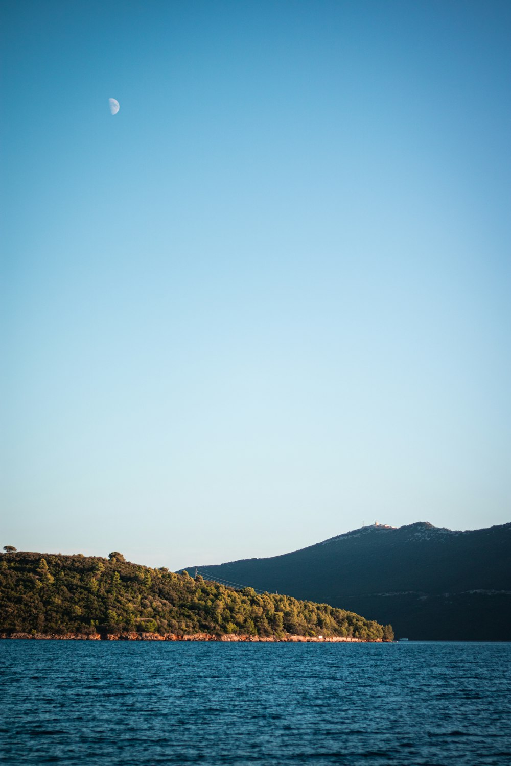 a body of water with a hill and a moon in the sky