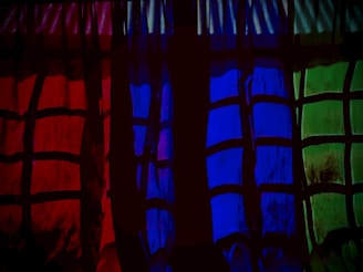 A series of curtains with a checkered pattern is illuminated in three distinct colors: red, blue, and green. The pattern consists of dark intersecting lines, creating blocks of varying shades. The ambient lighting accentuates the vibrant colors, casting a dramatic effect.