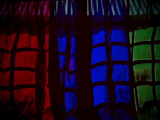 A series of curtains with a checkered pattern is illuminated in three distinct colors: red, blue, and green. The pattern consists of dark intersecting lines, creating blocks of varying shades. The ambient lighting accentuates the vibrant colors, casting a dramatic effect.
