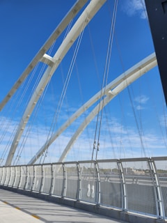 White arched suspender cables span across a modern, pedestrian bridge with tall, vertical supports against a vibrant blue sky. The bridge features a curved, perforated metal railing on one side, and the ground is made up of smooth concrete.