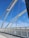 White arched suspender cables span across a modern, pedestrian bridge with tall, vertical supports against a vibrant blue sky. The bridge features a curved, perforated metal railing on one side, and the ground is made up of smooth concrete.