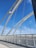 White arched suspender cables span across a modern, pedestrian bridge with tall, vertical supports against a vibrant blue sky. The bridge features a curved, perforated metal railing on one side, and the ground is made up of smooth concrete.