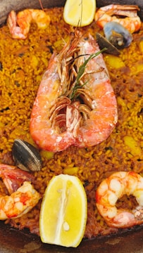 A close-up view of a seafood paella dish with yellow rice, large shrimp, mussels, and slices of lemon. The dish is rich in color and texture, featuring various seafood elements prominently placed on top of the seasoned rice.