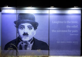A large mural on a wall depicts a monochrome portrait of a man with a bowler hat and a mustache, reminiscent of early 20th-century style. To the right, a quote reads: 'Laughter is the tonic, the relief, the surcease for pain. - Charlie Chaplin' alongside its Korean translation.