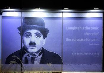 A large mural on a wall depicts a monochrome portrait of a man with a bowler hat and a mustache, reminiscent of early 20th-century style. To the right, a quote reads: 'Laughter is the tonic, the relief, the surcease for pain. - Charlie Chaplin' alongside its Korean translation.