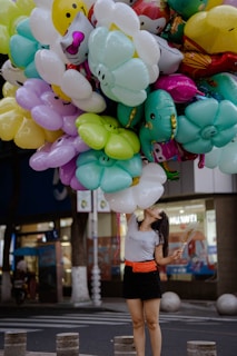 A person stands on a street holding a large bunch of colorful, cartoon-themed balloons. The balloons are primarily shaped like flowers and characters, with bright colors like yellow, green, pink, and blue. The person is wearing a gray t-shirt and a black skirt with an orange belt, looking upwards at the balloons. The background shows an urban setting with a storefront and trees.