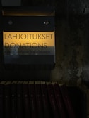 A donation box labeled in Finnish and English with the words 'Lahjoitukset Donations' and 'Kirkolle / For the Church'. The box is mounted on a textured wall. Below the box, several hymn books are neatly arranged in a row. The image is dimly lit, creating a somber and introspective atmosphere.
