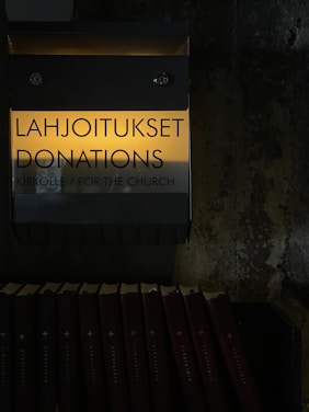 A donation box labeled in Finnish and English with the words 'Lahjoitukset Donations' and 'Kirkolle / For the Church'. The box is mounted on a textured wall. Below the box, several hymn books are neatly arranged in a row. The image is dimly lit, creating a somber and introspective atmosphere.