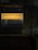 A donation box labeled in Finnish and English with the words 'Lahjoitukset Donations' and 'Kirkolle / For the Church'. The box is mounted on a textured wall. Below the box, several hymn books are neatly arranged in a row. The image is dimly lit, creating a somber and introspective atmosphere.