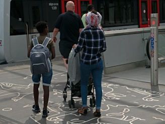 A group of people with bags and a stroller walking towards a modern tram. The ground features artistic line drawings, and the environment is urban with a modern public transit setting.