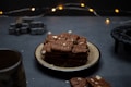 A stack of chocolate or gingerbread cookies is neatly arranged on a ceramic plate, surrounded by a dark, moody setting. In the background, there are blurred string lights that add a warm, festive touch. Cookie cutters are visible to the left, hinting at a baking theme.