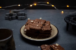 A stack of chocolate or gingerbread cookies is neatly arranged on a ceramic plate, surrounded by a dark, moody setting. In the background, there are blurred string lights that add a warm, festive touch. Cookie cutters are visible to the left, hinting at a baking theme.