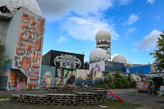 The image depicts an urban scene with a mix of street art and abandoned structures. Large murals cover the walls, featuring vibrant and eclectic designs, including faces and abstract patterns. In the background, there are spherical radar towers with a geodesic dome structure, hinting at a past military or scientific use. Wooden pallets are stacked in the foreground, resembling makeshift seating or decoration.