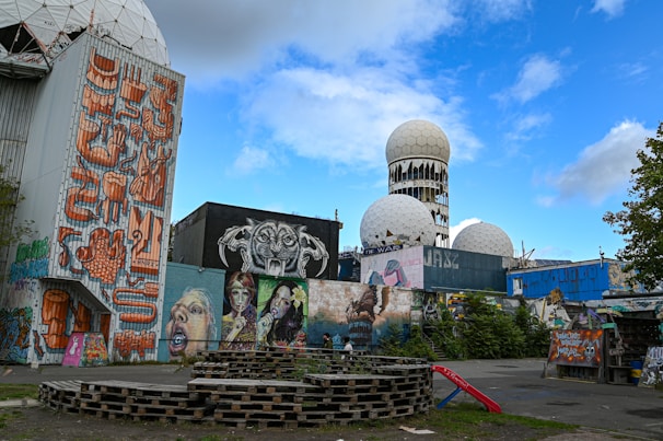 The image depicts an urban scene with a mix of street art and abandoned structures. Large murals cover the walls, featuring vibrant and eclectic designs, including faces and abstract patterns. In the background, there are spherical radar towers with a geodesic dome structure, hinting at a past military or scientific use. Wooden pallets are stacked in the foreground, resembling makeshift seating or decoration.
