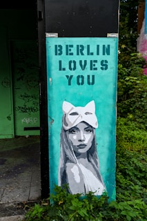 A mural on an urban structure depicts a woman with long hair wearing an animal-shaped mask. Above her, the text 'BERLIN LOVES YOU' is stenciled in bold capital letters. The mural is painted on a teal background and is partially surrounded by green foliage. Behind the mural, there is an area with graffiti on a door and walls.