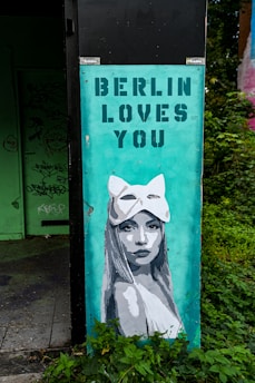 A mural on an urban structure depicts a woman with long hair wearing an animal-shaped mask. Above her, the text 'BERLIN LOVES YOU' is stenciled in bold capital letters. The mural is painted on a teal background and is partially surrounded by green foliage. Behind the mural, there is an area with graffiti on a door and walls.