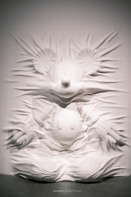 A textured wall sculpture features a prominent cartoon character with large round ears, a button nose, and gloved hands emerging from the surface. The material creates a flowing, fabric-like effect, adding depth and movement to the image.