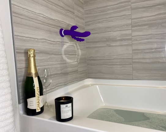 A bathroom scene with a bathtub partially filled with water. A bottle of Chandon champagne, an empty wine glass, and a black candle labeled 'V for Vibes' are placed on the corner ledge. The tiles are a gray marble pattern, and a purple suction cup mounted device is attached to the wall.