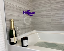 A bathroom scene with a bathtub partially filled with water. A bottle of Chandon champagne, an empty wine glass, and a black candle labeled 'V for Vibes' are placed on the corner ledge. The tiles are a gray marble pattern, and a purple suction cup mounted device is attached to the wall.