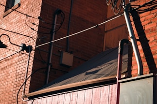 A brick wall with several black wires attached, and a metal roof at a diagonal angle. The wall features exposed electrical components and a window with metal bars. Shadows cast by the sun create patterns on the brick surface.