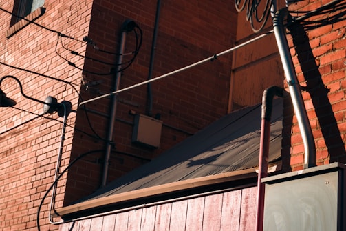 A brick wall with several black wires attached, and a metal roof at a diagonal angle. The wall features exposed electrical components and a window with metal bars. Shadows cast by the sun create patterns on the brick surface.